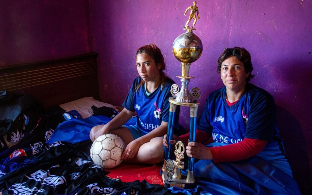 imagem de duas mulheres em um quarto, uma segur a bola a outra um trofeu e elas vestem camisas azuis