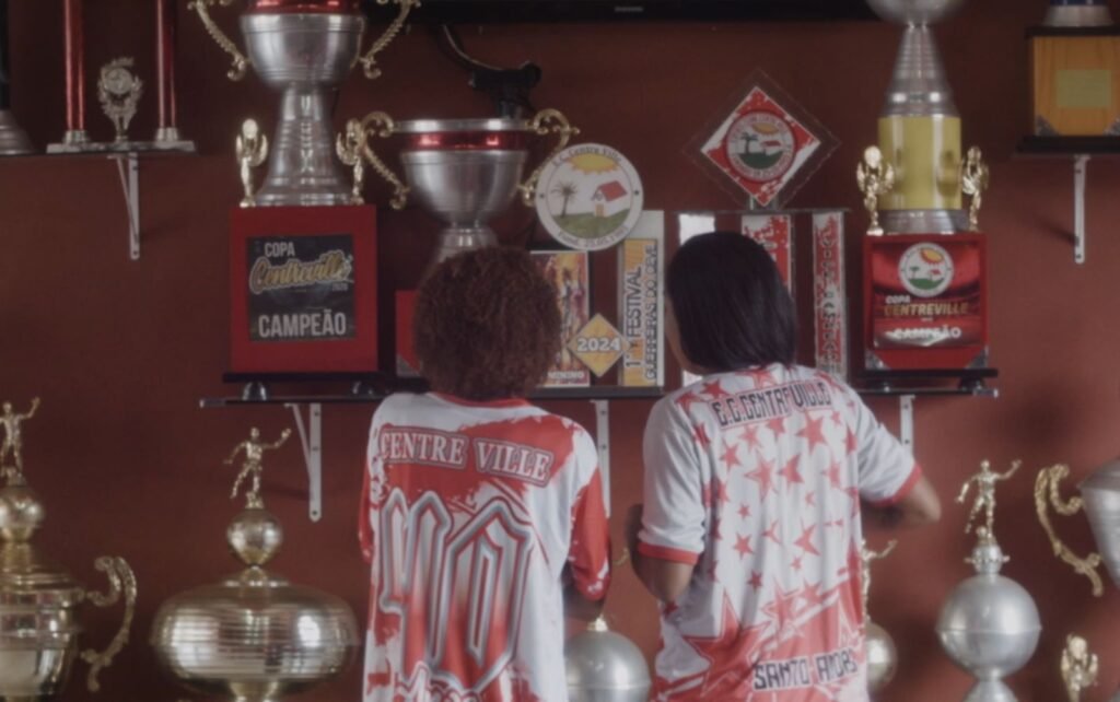 foto de duas mulheres de cstas, elas vestem camisa de time de varzea vermelho ebranco e olham para trofeus
