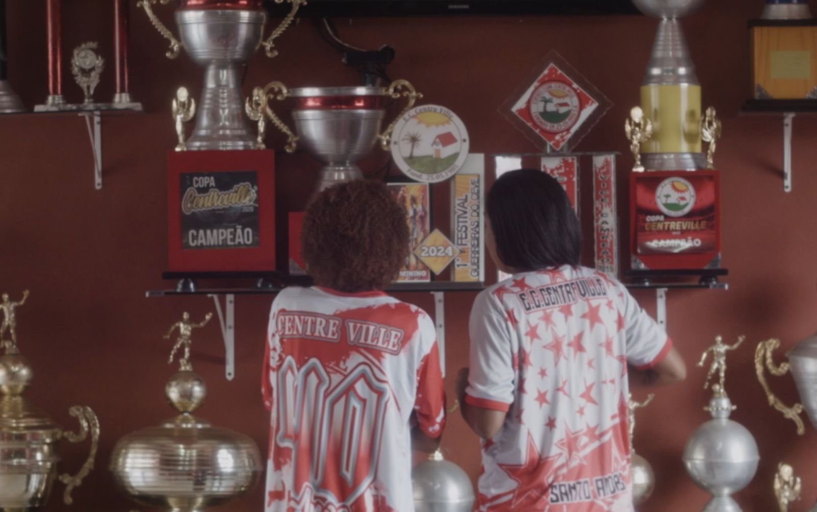 foto de duas mulheres de cstas, elas vestem camisa de time de varzea vermelho ebranco e olham para trofeus