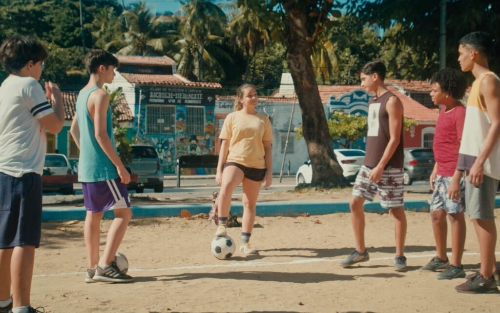 umagem de uma garota com o pé na bola rodeada de cinco garotos.