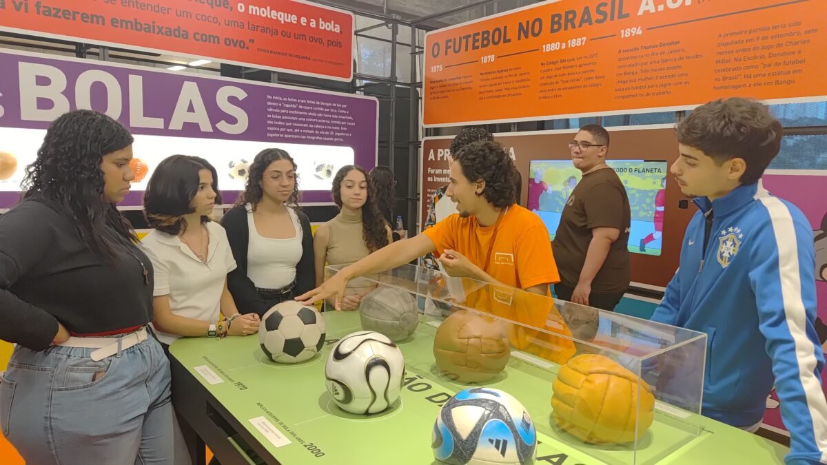 Um grupo de jovens escuta um educador do Museu apresentando uma coleção de bolas de futebol colocadas em uma mesa verde, que fica no meio da exposição principal, em um ambiente muito colorido.São moças e rapazes de perfis variados. O educado toca uma das bolas com a mão.
