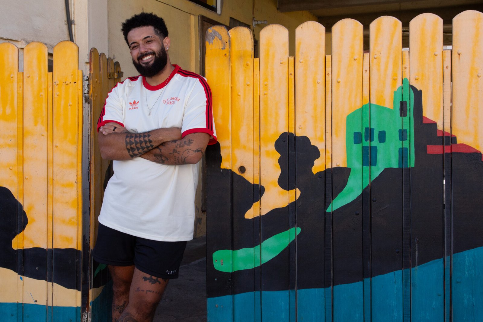 Foto do artista João Molambo encostado em um muro pintado por ele. Ele está à esquerda da foto de braços cruzados.