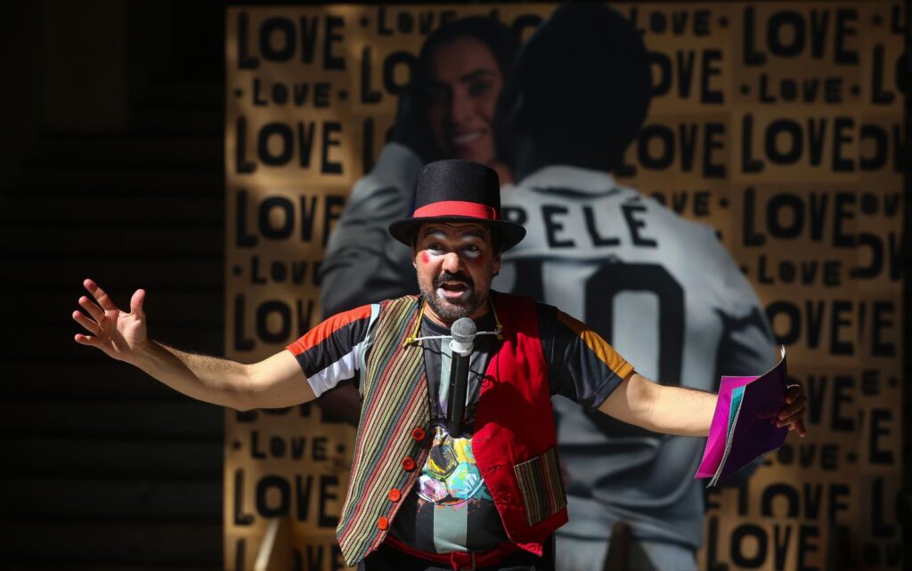 Imagem de um ator vestido de roupas coloridas e maquiagem. Ele está de braços abertos. Ao fundo, uma imagem da Marta e Pelé