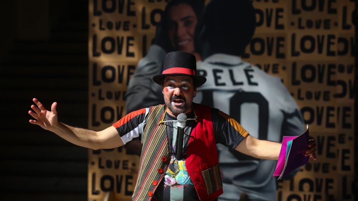 Imagem de um ator vestido de roupas coloridas e maquiagem. Ele está de braços abertos. Ao fundo, uma imagem da Marta e Pelé