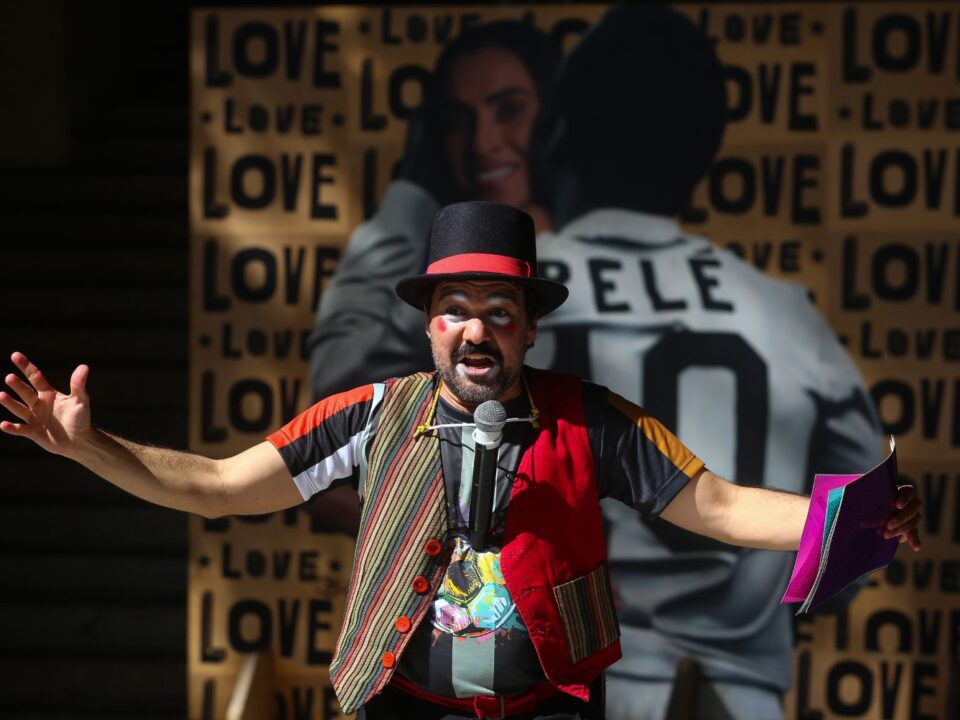 Imagem de um ator vestido de roupas coloridas e maquiagem. Ele está de braços abertos. Ao fundo, uma imagem da Marta e Pelé