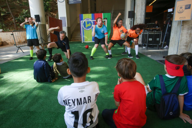 Imagem registrada na área externa do Museu do Futebol. Nela, crianças de costas assistem a uma peça de teatro.