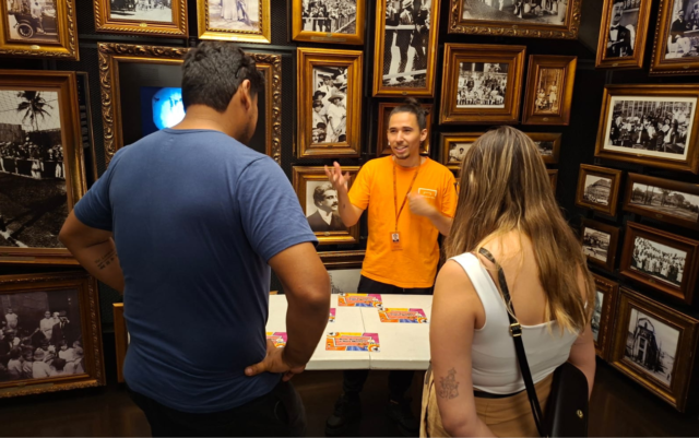 Foto da ativiade na Sala Origens; Educador está a frente de dois visitantes, realizando a atividade.
