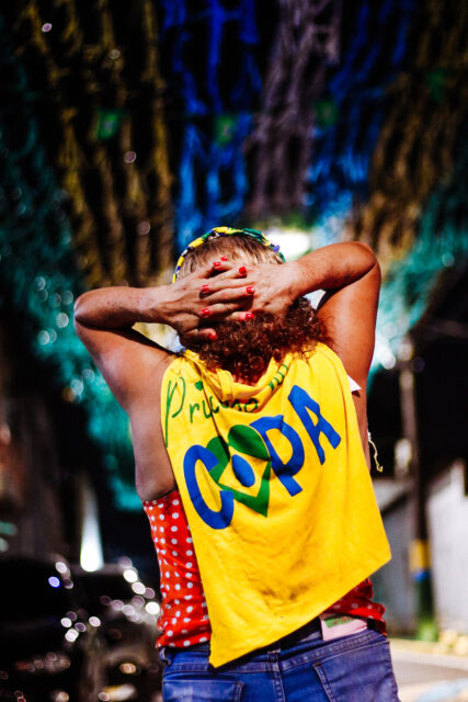 Uma mulher de costas com as mãos sobre as cabeças, as unhas pintadas de vermelho, e uma camiseta amarela com a palavra "copa" jogada sobre os ombros. Acima dela se vê fitas verdes, azuis e amarelos penduradas nos postes atravessando a rua.