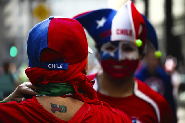 Dois torcedores do chile usam roupas e adereços azuis, vermelhos e amarelos. Nas coisas de um deles há uma tatuagem que mistura as bandeiras do Chile e do Brasil.