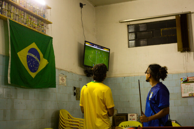 Em um bar, dois homens pausam o jogo de sinuca para ver um jogo de futebol na TV. Um veste a camisa amarela e outro a azul da seleção. À esquerda há uma bandeira do Brasil e à direita um calendário de 2026 pendurados na parede.
