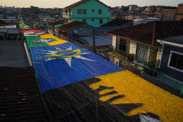 Uma rua de casas, se vê até o fim da cena desenhos formados com fitas coloridas penduradas, todos alusivos à seleção Brasileira de futebol.