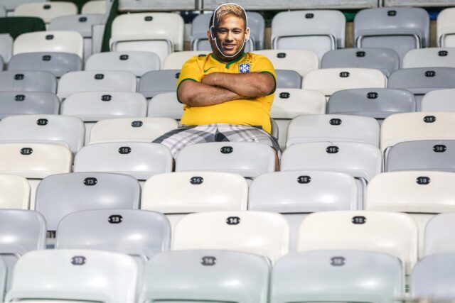 Num estádio vazio, com cadeiras brancas, um homem corpulento veste a camisa da seleção e uma máscara de papel com o rosto de Neyar. Ele está com os braços cruzados.