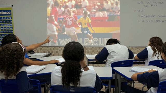 Numa sala de aula, está um grupo de meninas de costas para a câmera. elasolham para uma imagem da jogadora Sissi, com a camisa amarela da Seleção, driblando uma jogadora que veste vermelho. Uma das estudantes aponta para a tela.