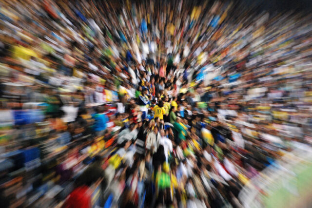 Torcida em um estádio de futebol. Com um efeito da fotografia, todos aparecem borrados, exceto uma pessoa focalizada de costas, com a camisa amarela 10 da seleção.