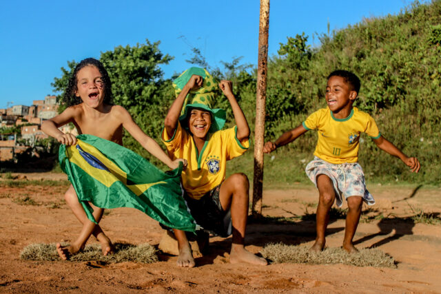 Três crianças em um caminho de terra riem como se estivessem comemorando algo. Uma delsa está sem camisa e tem nas mãos uma bandeira do Brasil. O do meio veste a camisa amarela da seleção e um chapéu alto verde e amarelo. O terceiro garoto também veste uma camisa amarela da seleção, onde se vê o número 10.