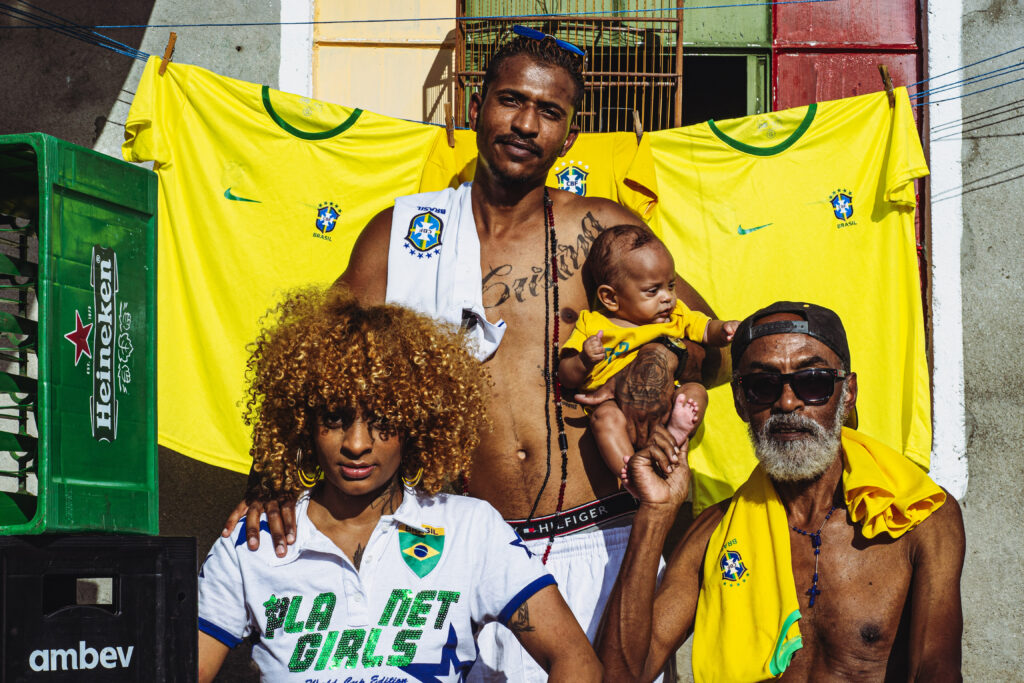 Retrato de uma família. Um homem de pé e sem camisa segura um bebê no colo, vestido de amarelo. Ele coloca a mão levemente no ombro de uma moça que está sentada à sua frente. Ela tem os cabelos loiros muito cacheados, na altura do queixo. Ela verste uma camiseta branca com um escudo do Brasil e os dizeres "Fla Net Girls" escrito em lantejoulas verdes. Ao lado dela, também sentado, está um senhor de barba branca, com uma camisa amarela do Brasil jogada sobre os ombros. ele usa um bon´preto virado para trás e óculos escuros, e segura com carinho o pé do bebê. No fundo da semana, há um varal com três camisas do Brasil. Do lado esquerdo da foto se vêem engradados de cerveja. Exceto o bebê, todos olham para a câmera.
