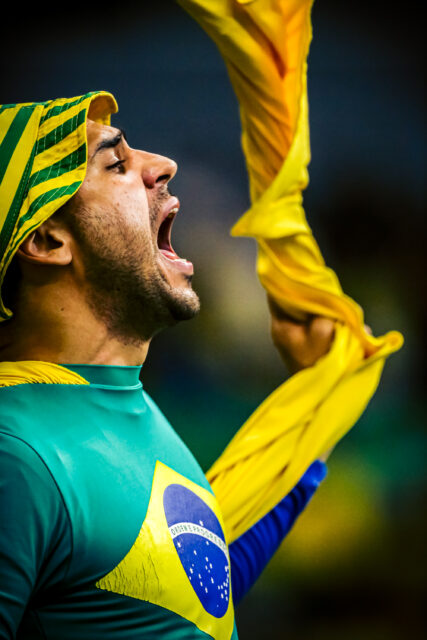 Homem branco, com barba por fazer, grita. Ele está vestido com uma camisa com a bandeira do Brasil e um gorro verde e amarelo. Na mão esquerda, gira um pano amarelo.