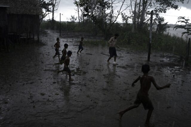 Cinco crianças e um homem adulto jogam bola na lama, durante uma chuva. A imagem tem tons escuros.