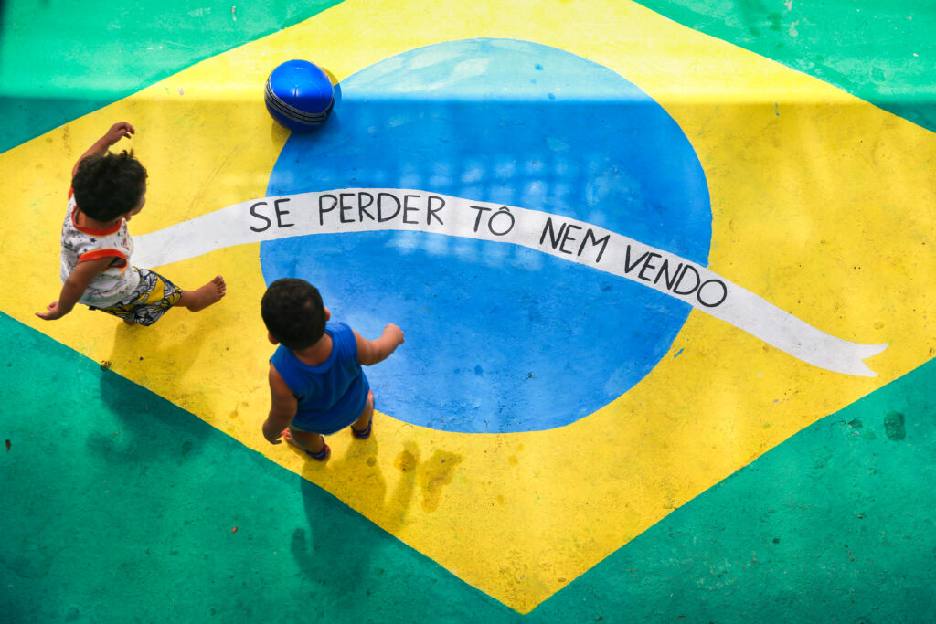 14 de Junho de 2018. Rua do bairro Pirambu decorada para a Copa do Mundo 2018. Uma bandeira do Brasil está pintada no chão e no lugar do "ordem e progresso" foi pintado "se perder tô nem vendo". Duas crianças peguenas jogam bola sobre a bandeira.