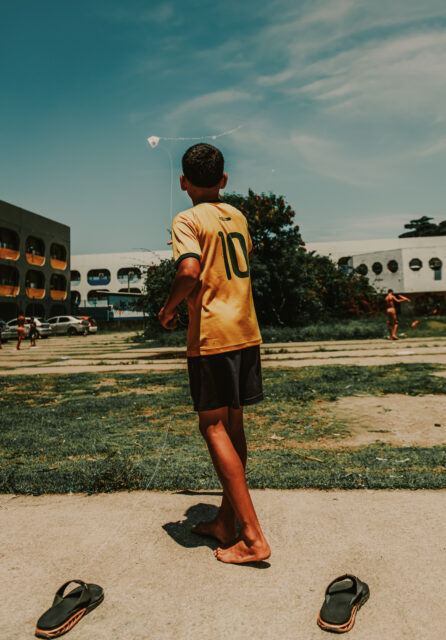 Um menino empina pipa com a camisa amarela 10 dda selação. Ele está descalço, mas seus chinelos estão logo atrás dele. No fundo, há edifícios do que parece ser uma grande escola.