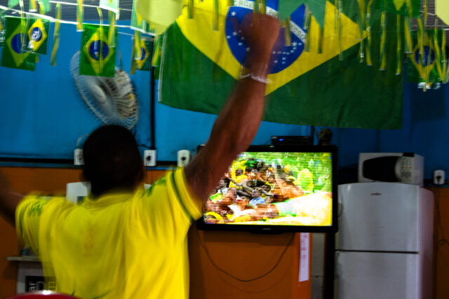 Um homem comemora sozinho em um bar, assistindo uma TV. Há várias bandeiras do Brasil penduradas no teto. O bar é modesto.