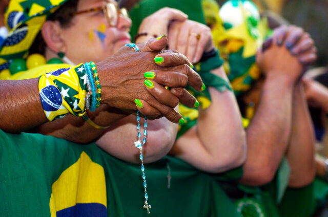 Torcida assiste jogo no Vale do Anhamgabaú durante o jogo BrasilX Mexico na Copa das confederaçoes de 2013. A imagem foca as mãos juntas em forma de oração. Em primeiro plano, as unhas são pintadas de verde e amarelo e seguram um terço de contas azuis.