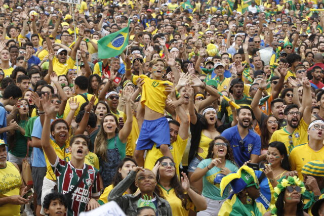 Multidão assistindo um jogo, quase todo mundo com a camisa do Brasil. Em destaque, um menino com o rosto pintado é levantado nos braços por um homem.