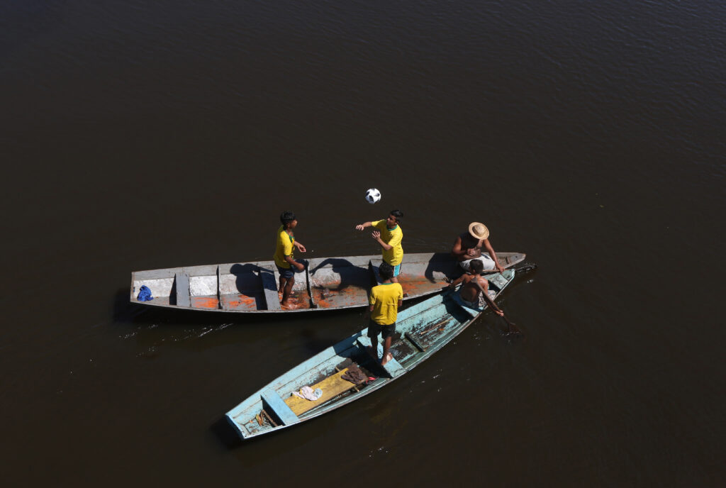Ribeirinhos com a bola oficial Telstar da Copa da Rússia de 2018 no Igarapé Açu em Manacapuru Municipio do Amazonas. Eles estão em canoas e são fotografados de cima. Três deles jogam bola com a camisa do Brasil. Outros dois esteão sentados e parecem conversar à frente dos barcos.