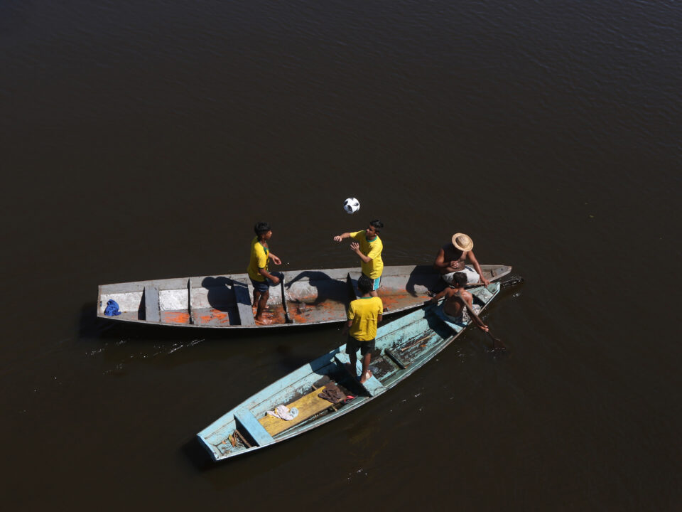 Ribeirinhos com a bola oficial Telstar da Copa da Rússia de 2018 no Igarapé Açu em Manacapuru Municipio do Amazonas. Eles estão em canoas e são fotografados de cima. Três deles jogam bola com a camisa do Brasil. Outros dois esteão sentados e parecem conversar à frente dos barcos.
