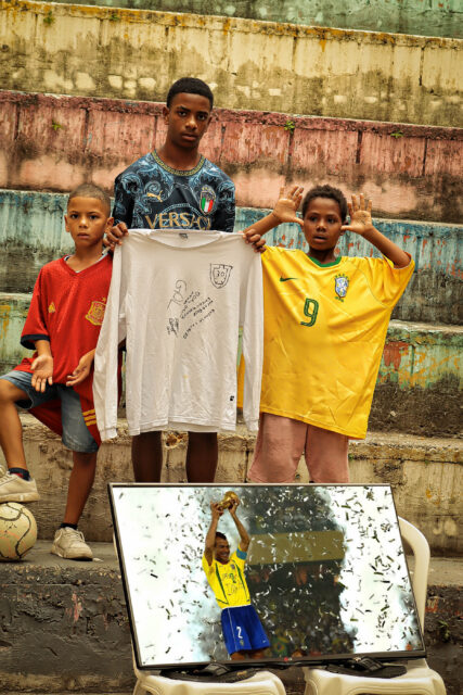 Três garotos posam para a foto em uma arquibancada de concreto, colorida, vestindo camisas de seleções: espanha, itália e Brasil. Aos pés deles, há uma fotografia do jogador Cafu levantando a taça. O que está com a camisa da Itália segura uma camiseta branca de mangas compridas onde alguém desenhou de caneta prta um escudo da CBF.