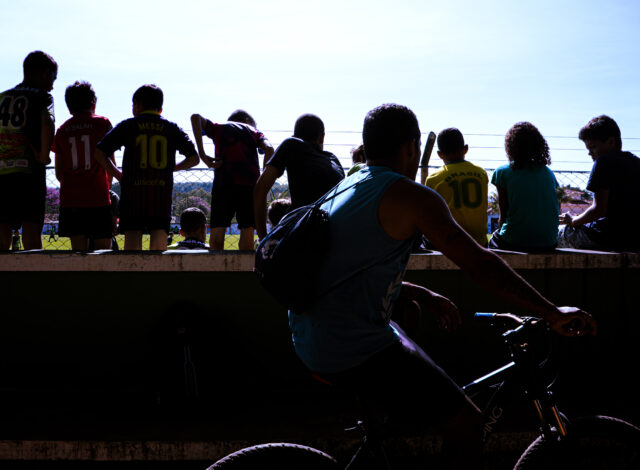 Um rgupo de crianças em contraluz assiste um jogo em um pequeno estádio. Em primeiro plano, há um homem de bicicleta. Uma das crianças veste a camisa amarela com o número 10 e a palavra Brasil