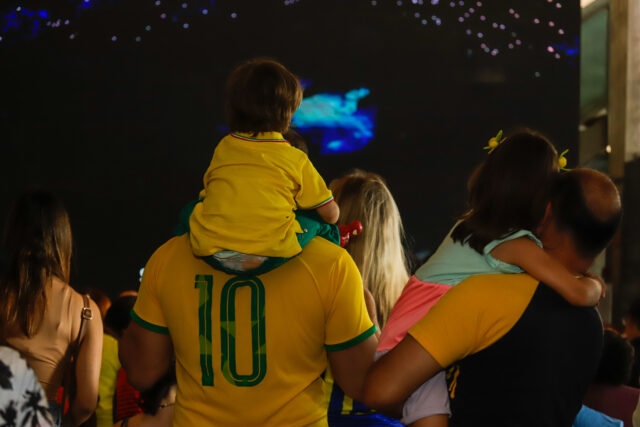 Pessoas assistem ao jogo do Brasil. Uma criança está com o filho em cima dos ombros. Ambos vestem a Amarelinha
