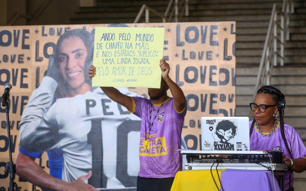 foto de uma das apresentações culturais já relaizadas no museu do Futebol. Em um painel com Marta e Pelé ao fundo, duas pessoas estão na foto. UMa esta sentada co a mesa de som e o ouyro, em pé, segura um cartaz com uma citação de Eduardo Galeano.