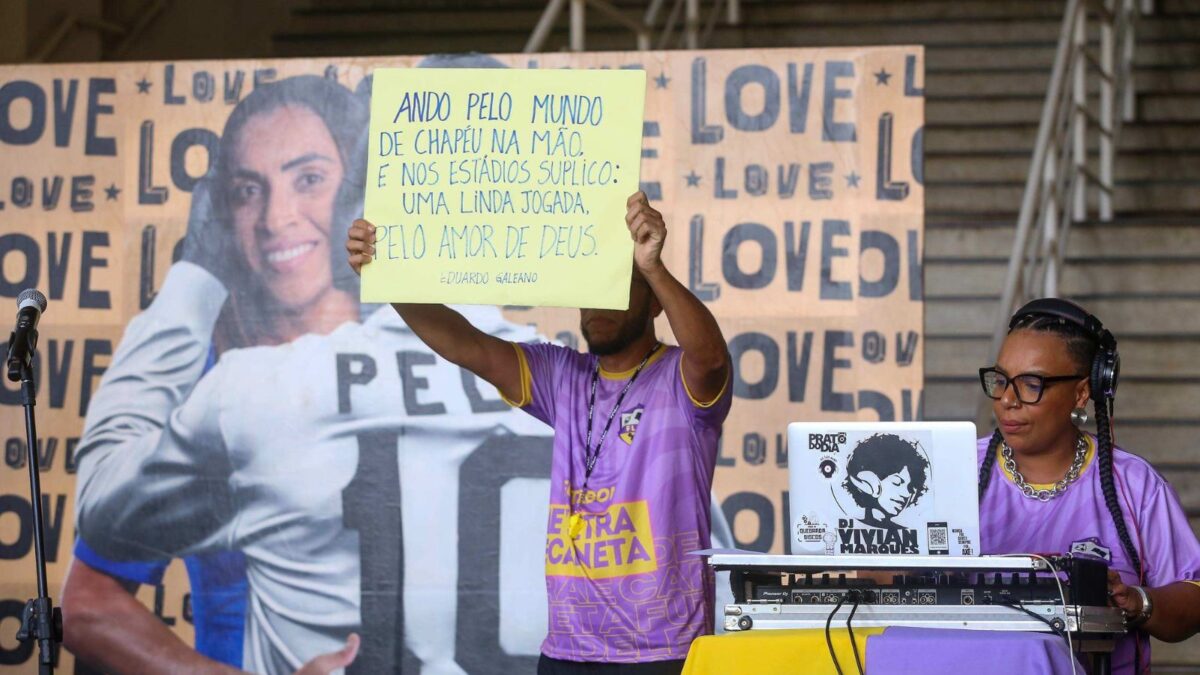 foto de uma das apresentações culturais já relaizadas no museu do Futebol. Em um painel com Marta e Pelé ao fundo, duas pessoas estão na foto. UMa esta sentada co a mesa de som e o ouyro, em pé, segura um cartaz com uma citação de Eduardo Galeano.