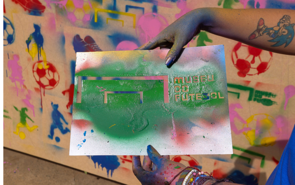 imagem de um muro pintado em grafite. uma mão segura um desenho em grafite e nele está escrito: Museu do Futebol.