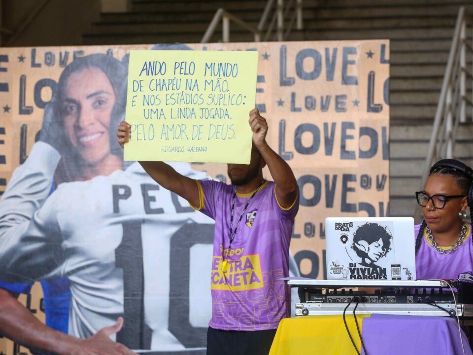 foto de uma das apresentações culturais já relaizadas no museu do Futebol. Em um painel com Marta e Pelé ao fundo, duas pessoas estão na foto. UMa esta sentada co a mesa de som e o ouyro, em pé, segura um cartaz com uma citação de Eduardo Galeano.