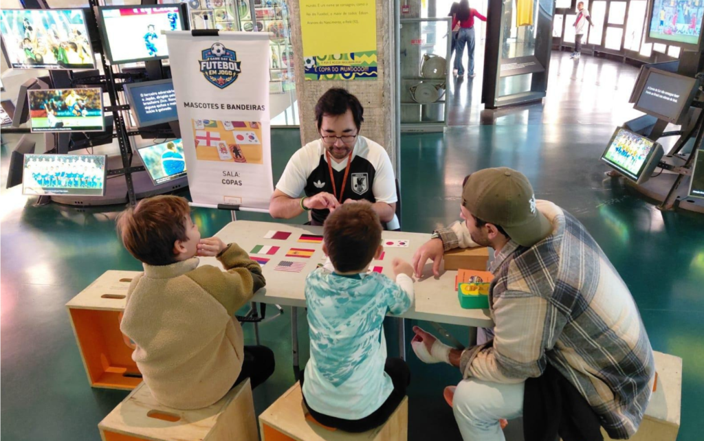 Foto na sala das Copas no Game Day anterior. Nele um educador está sentando em uma mesa, com os viistantes de costas, fazendo uma das atividades promovidas pelo educativo do museu