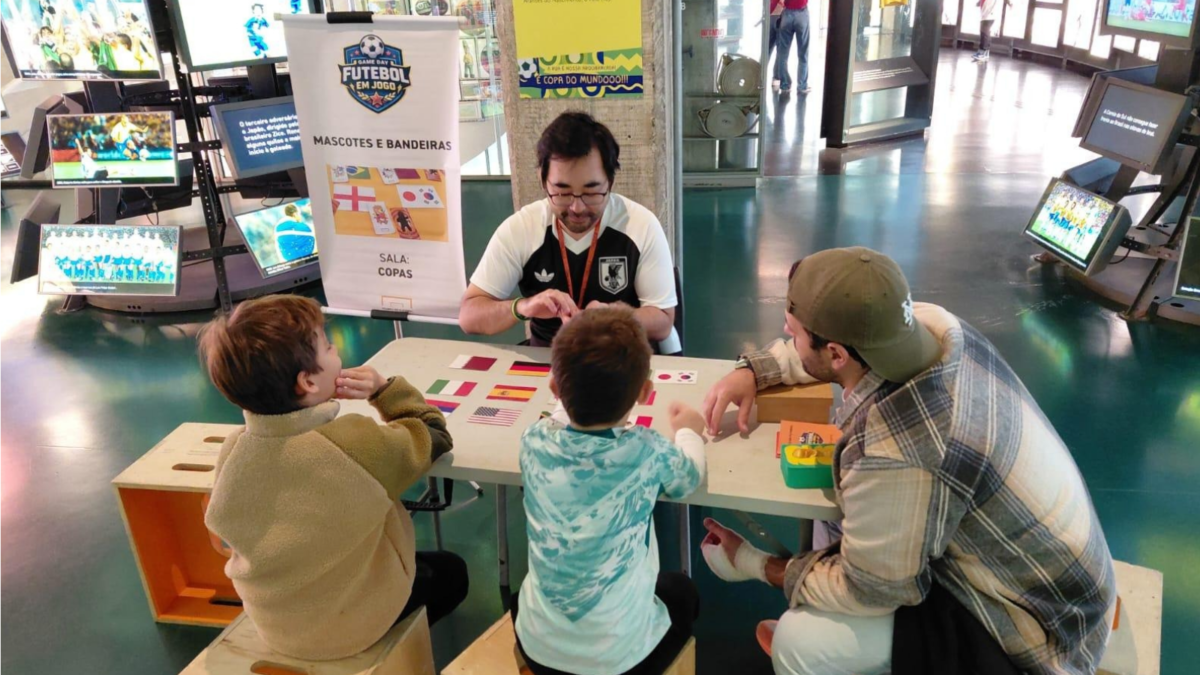 Foto na sala das Copas no Game Day anterior. Nele um educador está sentando em uma mesa, com os viistantes de costas, fazendo uma das atividades promovidas pelo educativo do museu