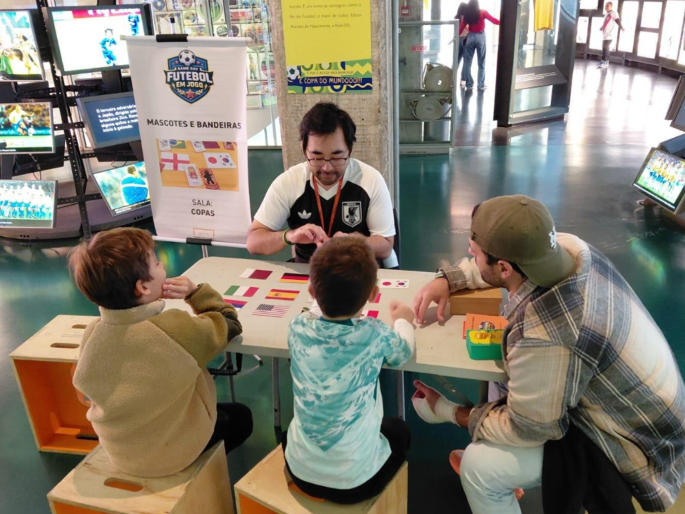 Foto na sala das Copas no Game Day anterior. Nele um educador está sentando em uma mesa, com os viistantes de costas, fazendo uma das atividades promovidas pelo educativo do museu